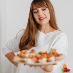 Smiling woman in a white top holding a plate of fresh canapés in a modern kitchen setting.