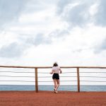 A woman stands on a red sand cliff overlooking the ocean, under a cloudy sky in Natal, Brazil.